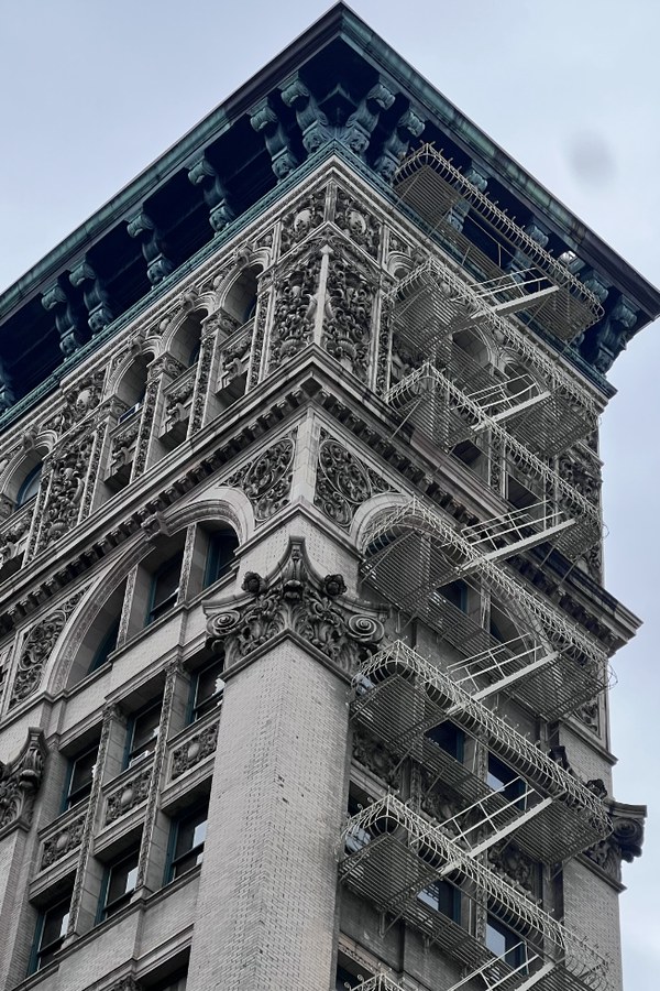 Upward view of an ornate building corner with a green-blue cornice and fire escapes
